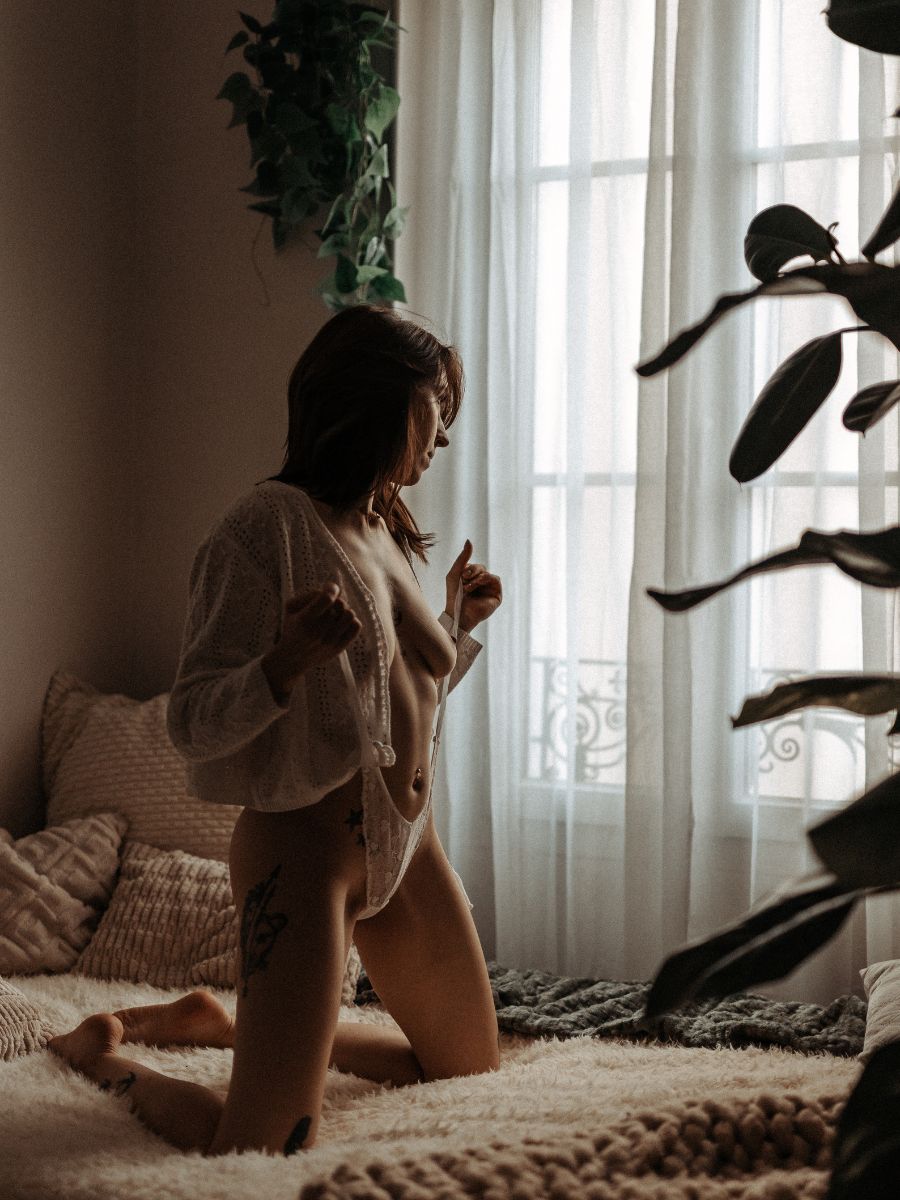 Portrait boudoir à genoux sur un lit, cardigan blanc entrouvert devant une fenêtre.
