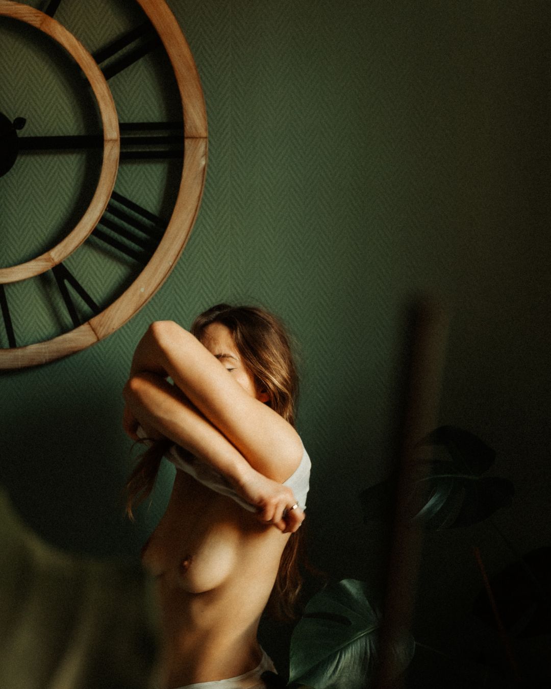 Portrait boudoir de profil, bras replié sur le visage, buste dénudé près d'une horloge.